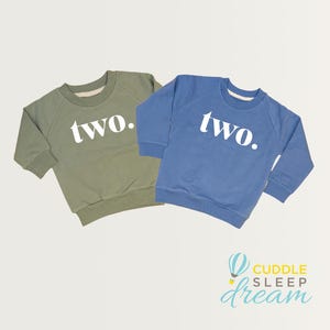 May include: Two sweatshirts, one olive green and one blue, are displayed. Each has the word "two." printed in white across the front. The sweatshirts are likely for a toddler's second birthday. The Cuddle Sleep Dream logo is in the bottom right corner.
