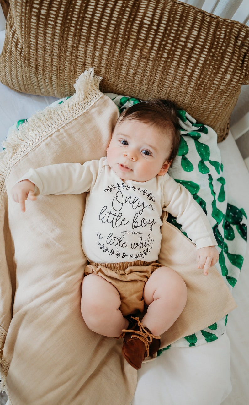 Pode incluir: Um beb&ecirc; vestindo um macac&atilde;o branco com o texto "Only a little boy - for such a little while" em letras pretas. O beb&ecirc; est&aacute; deitado em um cobertor bege com um cobertor estampado verde e branco por baixo.