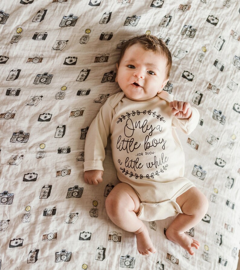 Pode incluir: Um beb&ecirc; vestindo um macac&atilde;o branco com texto preto que diz "Only a little boy - for such a little while". O macac&atilde;o &eacute; decorado com pequenos desenhos florais. O beb&ecirc; est&aacute; deitado em um cobertor branco com um padr&atilde;o de c&acirc;meras pretas.