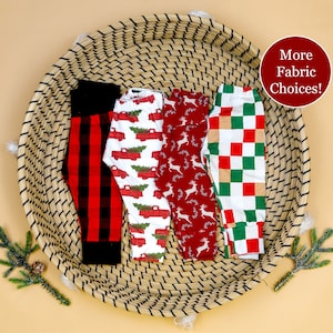 May include: Four pairs of red and white patterned leggings in a woven basket. The patterns include plaid, reindeer, Christmas trees, and trucks. The text "More Fabric Choices!" is in a red circle.