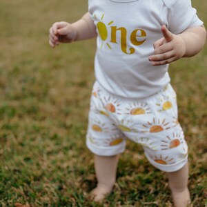 May include: A toddler wearing a white onesie with a yellow sun and the word "one" printed on it. The toddler is wearing white shorts with a pattern of yellow suns. The toddler is standing on green grass.