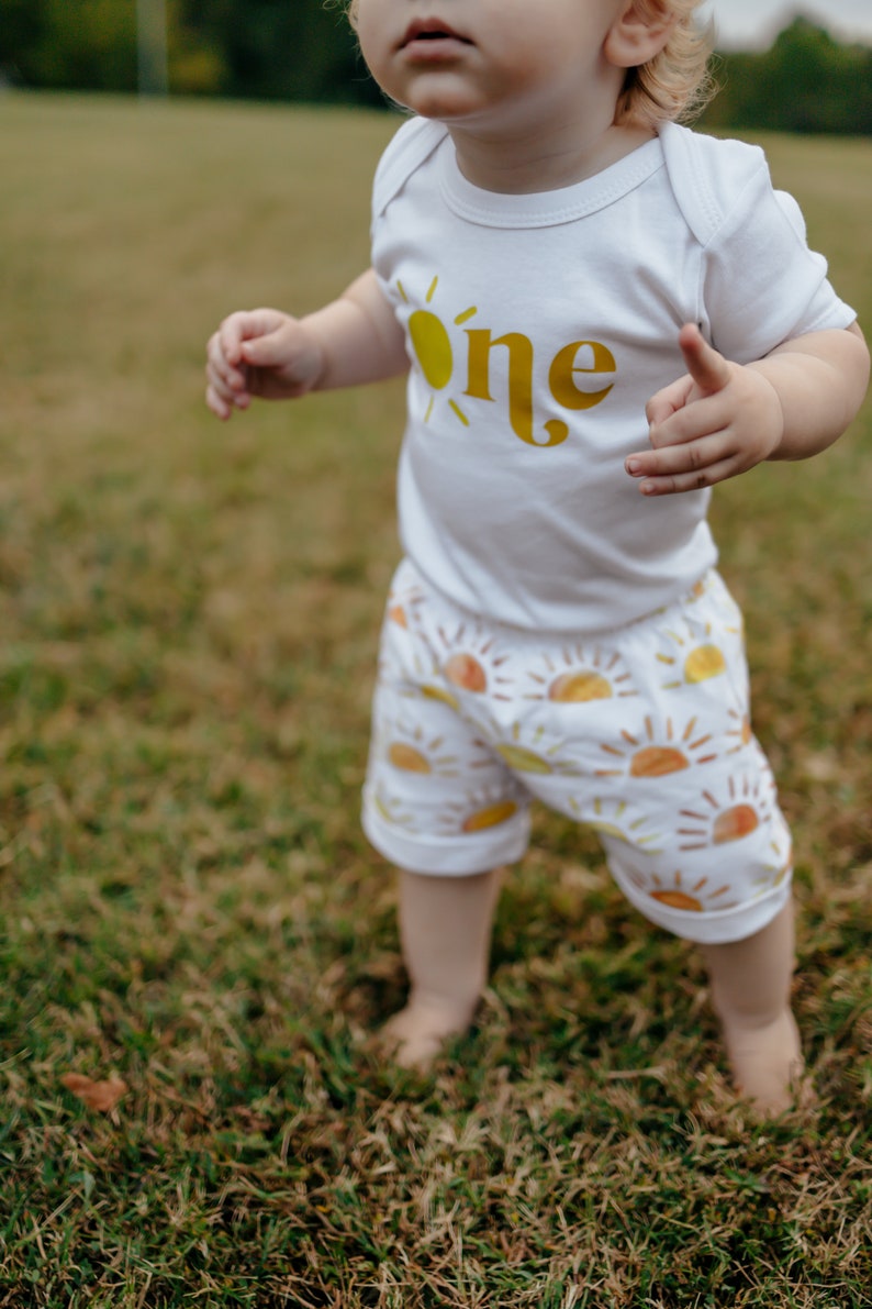 May include: A toddler wearing a white onesie with a yellow sun and the word "one" printed on it. The toddler is wearing white shorts with a pattern of yellow suns. The toddler is standing on green grass.