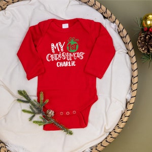 May include: Red long-sleeve baby onesie with white text that reads "My 1st Christmas Charlie" and a green and red Christmas ornament graphic.