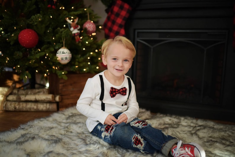May include: A young boy wearing a white long-sleeved shirt with black suspenders and a red and black bow tie. He is wearing blue jeans with red and black patches and red and white sneakers. He is sitting on a white furry rug in front of a fireplace.