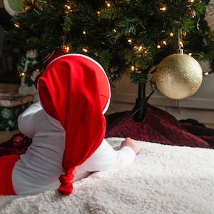 May include: A baby wearing a red and white Santa hat and a red and white onesie is lying on a white fluffy blanket. The baby is looking at a Christmas tree with gold baubles.
