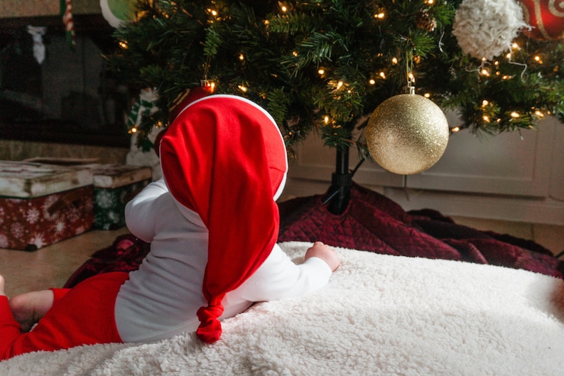 May include: A baby wearing a red and white Santa hat and a red and white onesie is lying on a white fluffy blanket. The baby is looking at a Christmas tree with gold baubles.