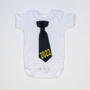May include: White baby onesie with a black tie applique. The tie has the gold glitter text "2022" on it. The onesie has short sleeves and three snaps at the bottom.