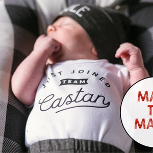 Puede incluir: Un body blanco con texto negro que dice "Just Joined Team Castan". Un beb&eacute; que lleva el body est&aacute; acostado sobre una superficie a cuadros negros y blancos. Un c&iacute;rculo rojo con el texto "Made to Match" est&aacute; en la esquina inferior derecha de la imagen.