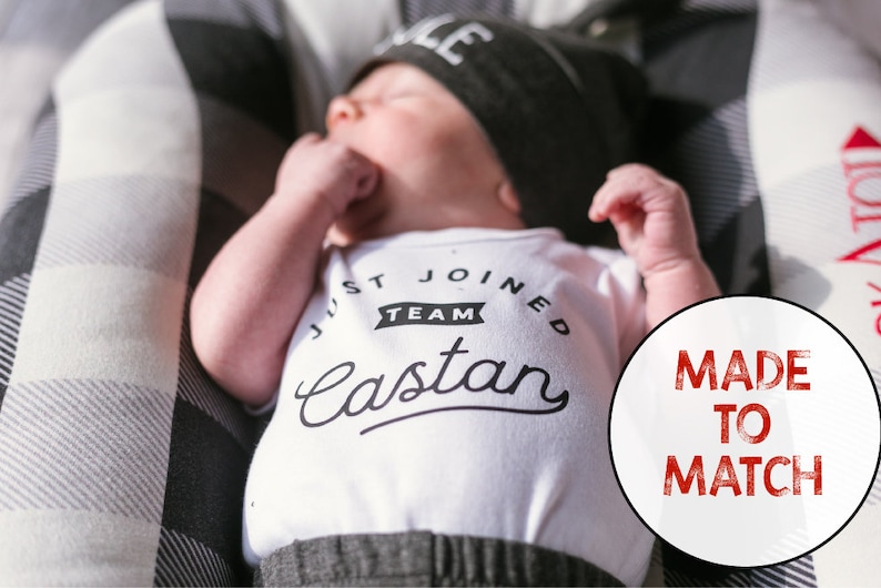 Puede incluir: Un body blanco con texto negro que dice "Just Joined Team Castan". Un beb&eacute; que lleva el body est&aacute; acostado sobre una superficie a cuadros negros y blancos. Un c&iacute;rculo rojo con el texto "Made to Match" est&aacute; en la esquina inferior derecha de la imagen.