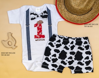 May include: A white baby bodysuit with blue suspenders, a black and white cow print bow tie, and matching shorts. The number "1" is red with a paisley design, and the name "Weston" is below it. A cowboy hat graphic is on the bodysuit.