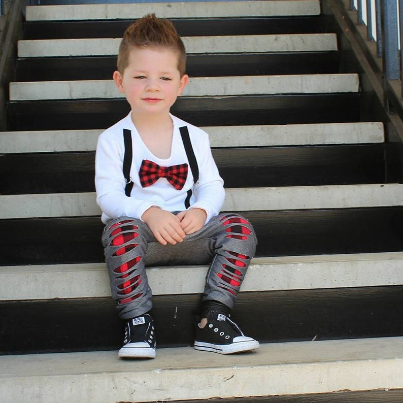 May include: A young boy wearing a white long-sleeved shirt with black suspenders and a red and black bow tie. He is wearing grey pants with red and black plaid patches and black sneakers. He is sitting on a set of stairs.