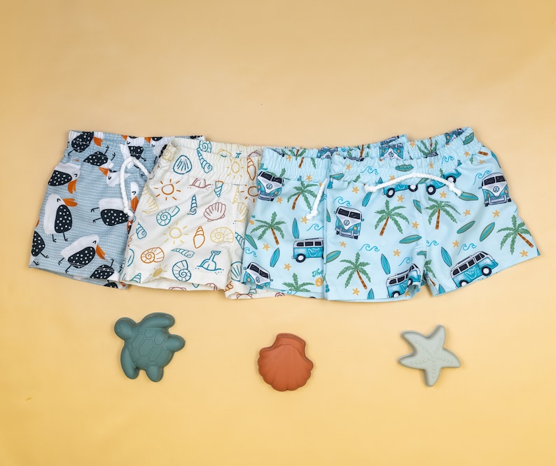 May include: Three pairs of colourful swimming trunks with different patterns. One pair is blue with white camper vans and palm trees, another is white with seashells and a sun, and the last is blue with white seagulls.