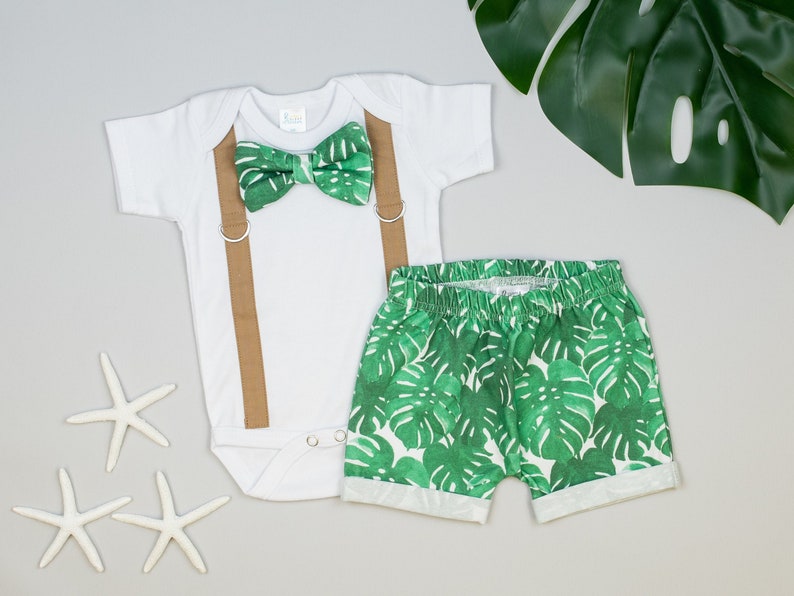 May include: A white bodysuit with brown suspenders and a green bow tie with a tropical leaf pattern. The outfit is paired with green shorts featuring a tropical leaf print.