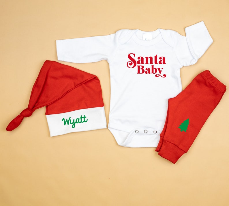 May include: A white long-sleeved onesie with the text "Santa Baby" in red. A red and white Santa hat with the name "Wyatt" embroidered in green. Red trousers with a green Christmas tree design.