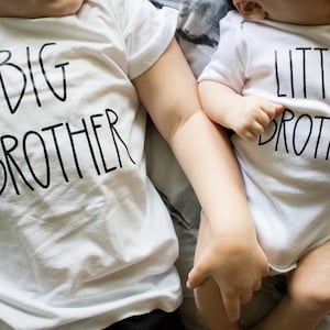 Op de afbeelding: Twee baby's dragen witte T-shirts met zwarte tekst. De oudere baby draagt een shirt met de tekst "GROTE BROER" en de jongere baby draagt een shirt met de tekst "KLEINE BROER".