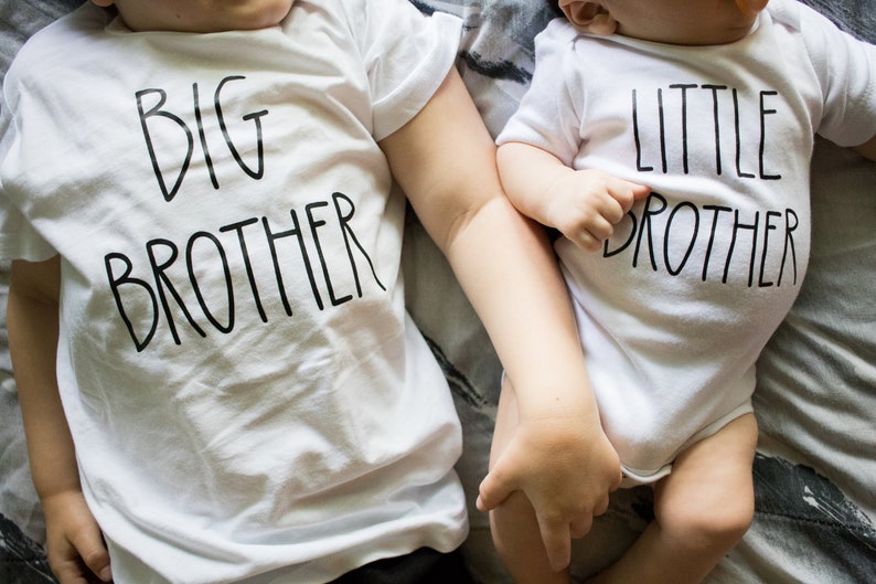 Op de afbeelding: Twee baby's dragen witte T-shirts met zwarte tekst. De oudere baby draagt een shirt met de tekst "GROTE BROER" en de jongere baby draagt een shirt met de tekst "KLEINE BROER".