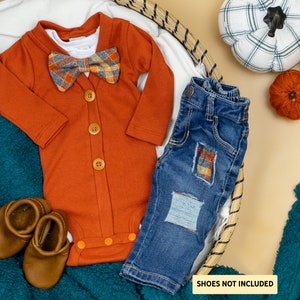 May include: A rust-colored baby onesie with a plaid bow tie, blue jeans with plaid patches, and brown baby shoes. The onesie has a button closure down the front. "Shoes not included" is written on the bottom right corner of the image.