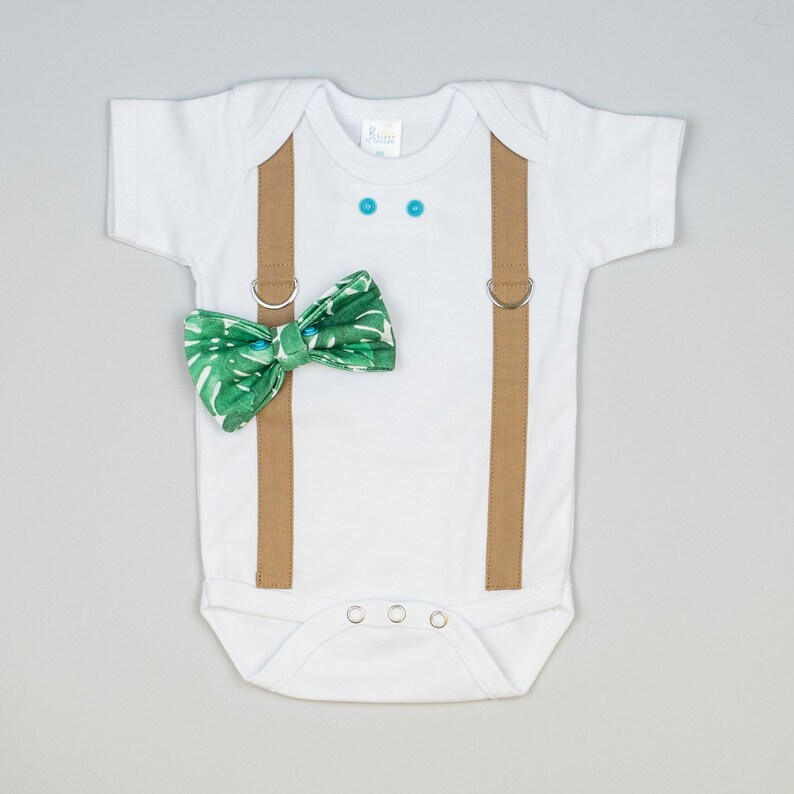 May include: A white baby onesie with brown suspenders and a green bow tie with a tropical leaf pattern. The onesie has two blue buttons on the chest and three silver snaps on the crotch.