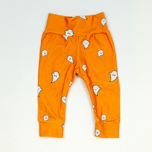 Pu&ograve; includere: Leggings arancioni con un motivo a fantasmi bianchi. I fantasmi hanno facce sorridenti.