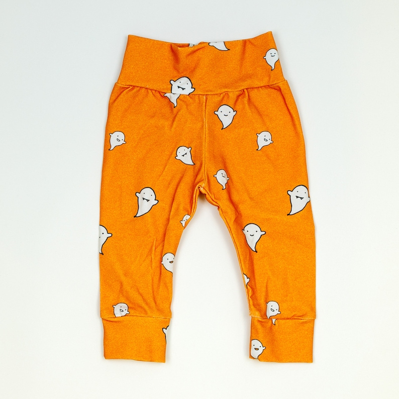 Pu&ograve; includere: Leggings arancioni con un motivo a fantasmi bianchi. I fantasmi hanno facce sorridenti.