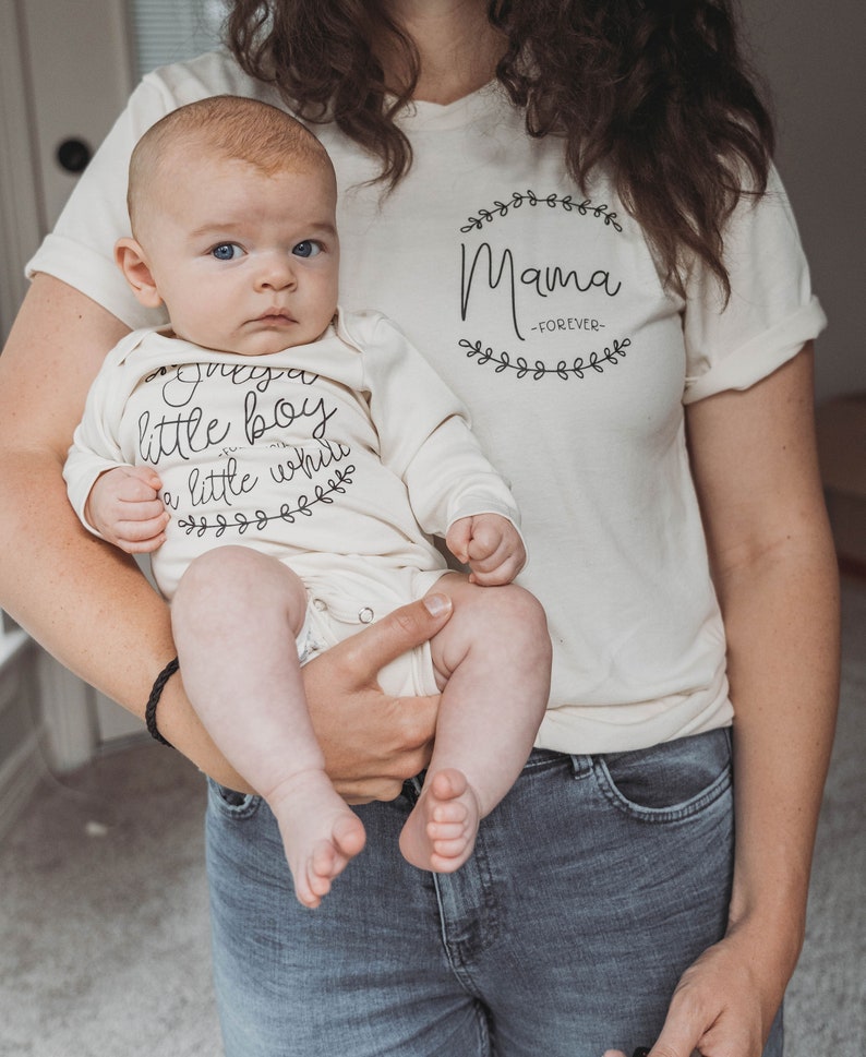 May include: A woman holding a baby wearing a white onesie with the text "Just a little boy with a little while" and a floral design. The woman is wearing a white t-shirt with the text "Mama -FOREVER- " and a floral design.