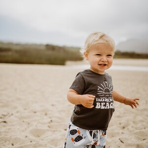 May include: A young child wearing a dark grey t-shirt with the text "Take me to the beach" and white and black patterned shorts. The child is standing on a sandy beach.
