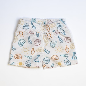 May include: White cotton shorts with a beach-themed print featuring seashells, suns, and shovels in shades of blue, orange, and brown.