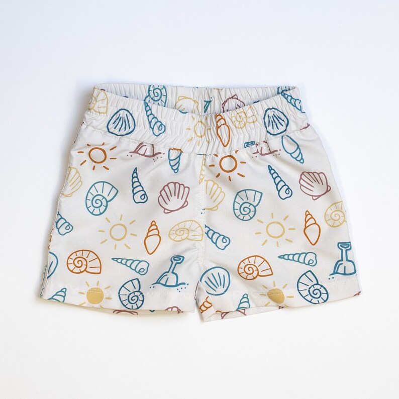 May include: White cotton shorts with a beach-themed print featuring seashells, suns, and shovels in shades of blue, orange, and brown.
