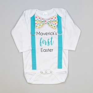 Puede incluir: Mono blanco de manga larga para beb&eacute; con tirantes turquesa y una pajarita de lunares multicolores. El mono tiene el texto "Maverick's first Easter" impreso.