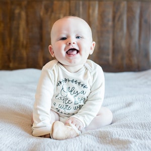 Pode incluir: Um beb&ecirc; vestindo um macac&atilde;o branco de manga comprida com texto preto que diz "Only a little boy a cute one". O beb&ecirc; est&aacute; sentado em uma colcha branca e sorrindo.
