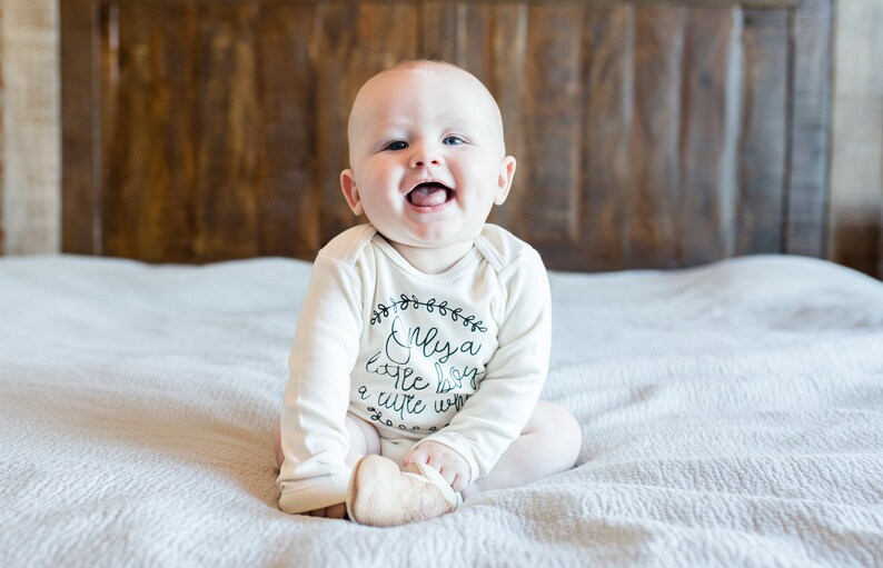 Pode incluir: Um beb&ecirc; vestindo um macac&atilde;o branco de manga comprida com texto preto que diz "Only a little boy a cute one". O beb&ecirc; est&aacute; sentado em uma colcha branca e sorrindo.