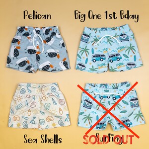 May include: Four pairs of toddler swim trunks with different patterns. The patterns include pelicans, sea shells, and a blue van with the words "The Big One" on it. One pair of swim trunks is marked "Sold Out".