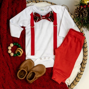 May include: A white baby onesie with red suspenders and a plaid bow tie. The onesie is laid out on a red knitted blanket with a pair of brown leather baby shoes and a wooden and green teething ring.