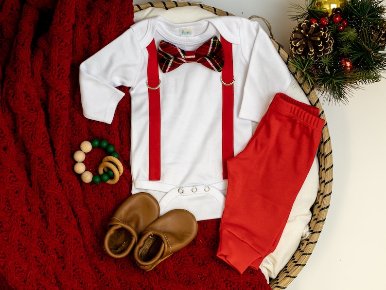 May include: A white baby onesie with red suspenders and a plaid bow tie. The onesie is laid out on a red knitted blanket with a pair of brown leather baby shoes and a wooden and green teething ring.