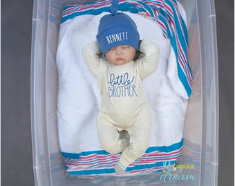 May include: A baby wearing a blue knitted hat with the name "BENNETT" embroidered on it and a cream coloured onesie with the words "little brother" in blue. The baby is lying on a white and blue striped blanket.