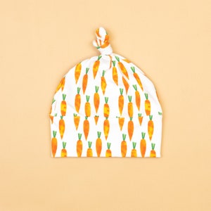 May include: A white baby hat featuring a repeating pattern of orange carrots with green tops. The hat is knotted at the top. The background is a plain light orange colour.
