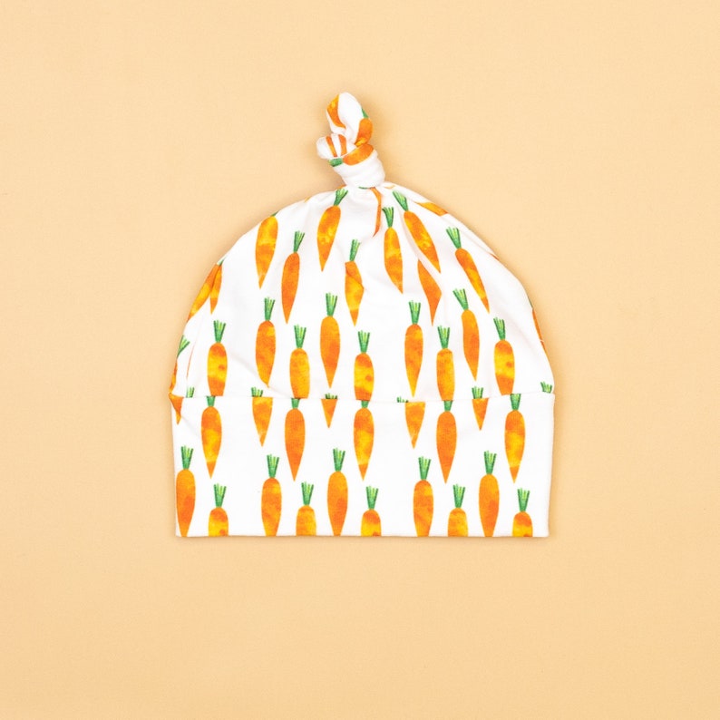 May include: A white baby hat featuring a repeating pattern of orange carrots with green tops. The hat is knotted at the top. The background is a plain light orange colour.