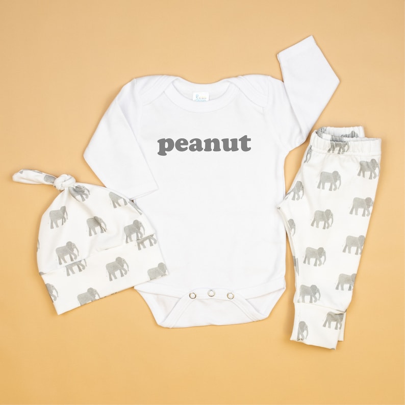 May include: A white baby outfit set with an elephant print. The set includes a long-sleeved bodysuit with the word "peanut" printed on the front, a matching hat, and leggings.