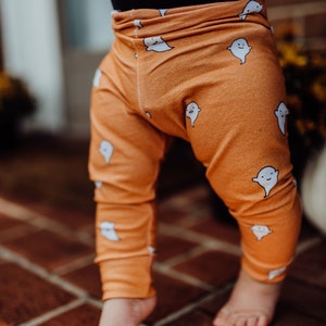 Pu&ograve; includere: Leggings arancioni con un motivo a fantasmi bianchi. I leggings sono indossati da un bambino in piedi su un pavimento di mattoni.