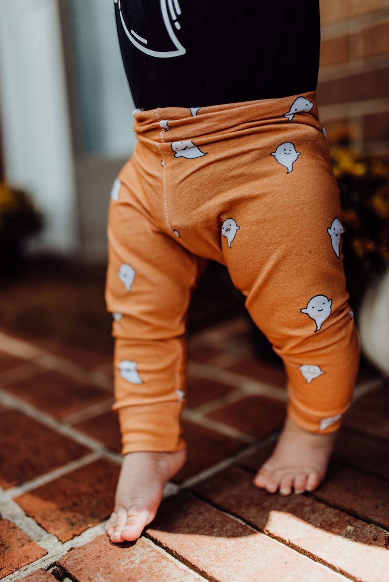 Pu&ograve; includere: Leggings arancioni con un motivo a fantasmi bianchi. I leggings sono indossati da un bambino in piedi su un pavimento di mattoni.