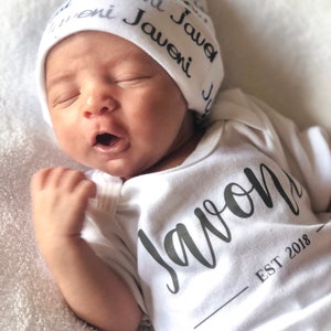 May include: A white baby onesie with the name "Javon" written in black cursive lettering and the text "EST 2018" below. The baby is wearing a white knit hat with the name "Javeni" repeated in black lettering.