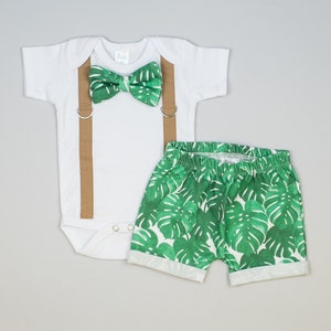 May include: A white bodysuit with brown suspenders and a green bow tie. The bodysuit is paired with green shorts featuring a tropical leaf print.