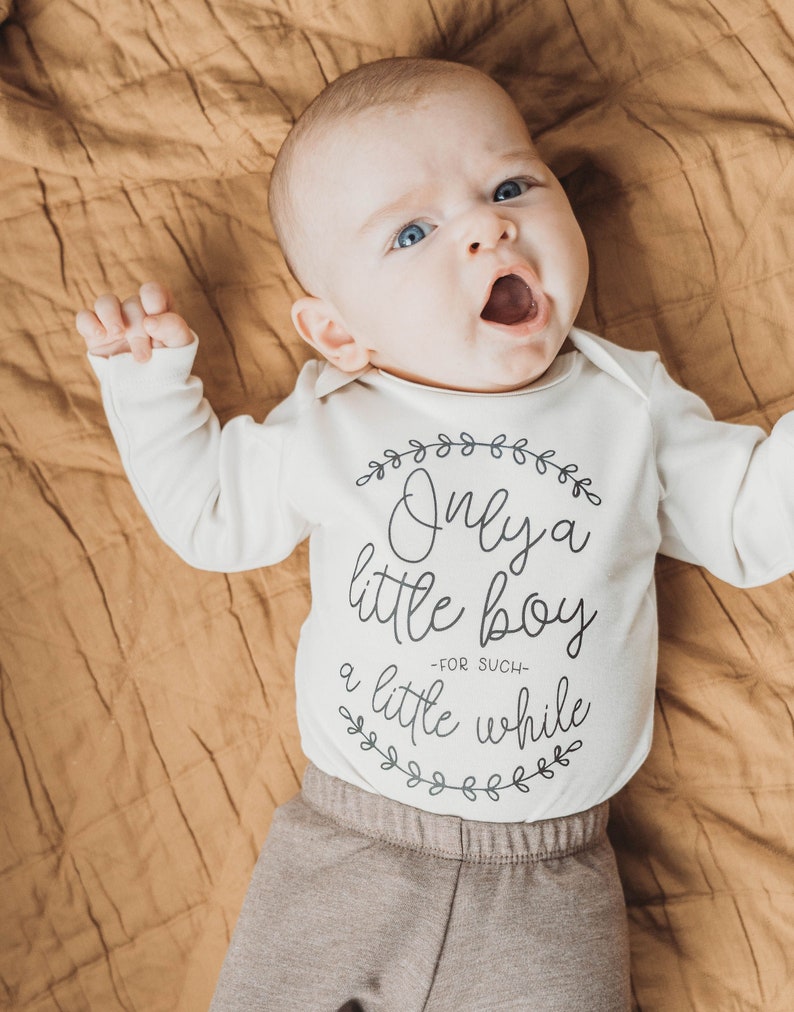 Pode incluir: Um macac&atilde;o branco para beb&ecirc; com texto preto que diz "Only a little boy -for such- a little while" cercado por uma coroa de folhas. O macac&atilde;o &eacute; usado por um beb&ecirc; deitado em um cobertor marrom.