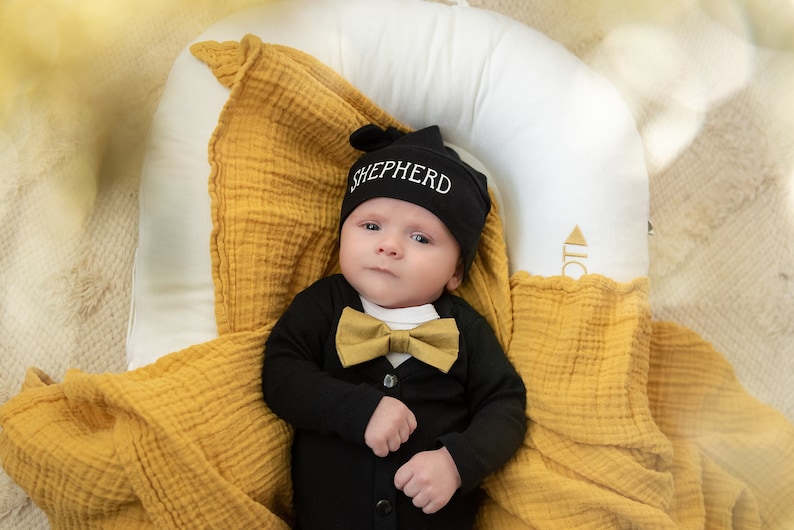 Puede incluir: Un beb&eacute; con un mono negro, una pajarita dorada y un gorro negro que dice "SHEPHERD". El beb&eacute; est&aacute; tumbado en una manta amarilla.