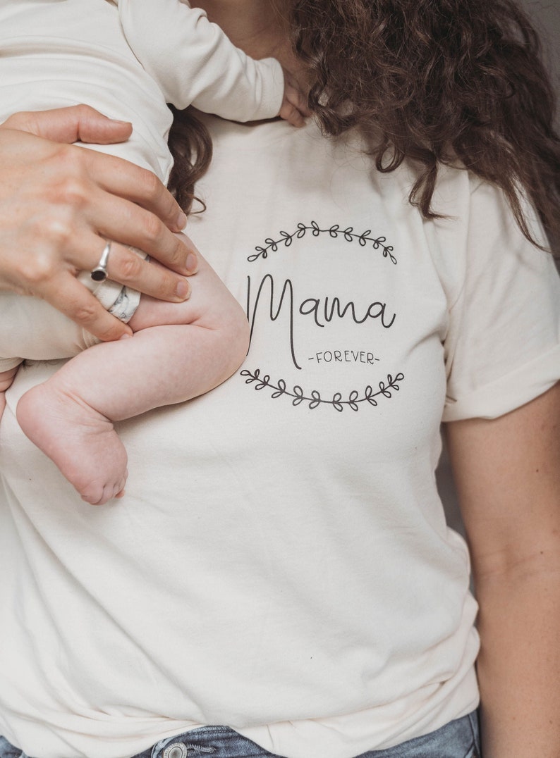 May include: A white t-shirt with a black graphic that says "Mama -FOREVER- " surrounded by a wreath of leaves.