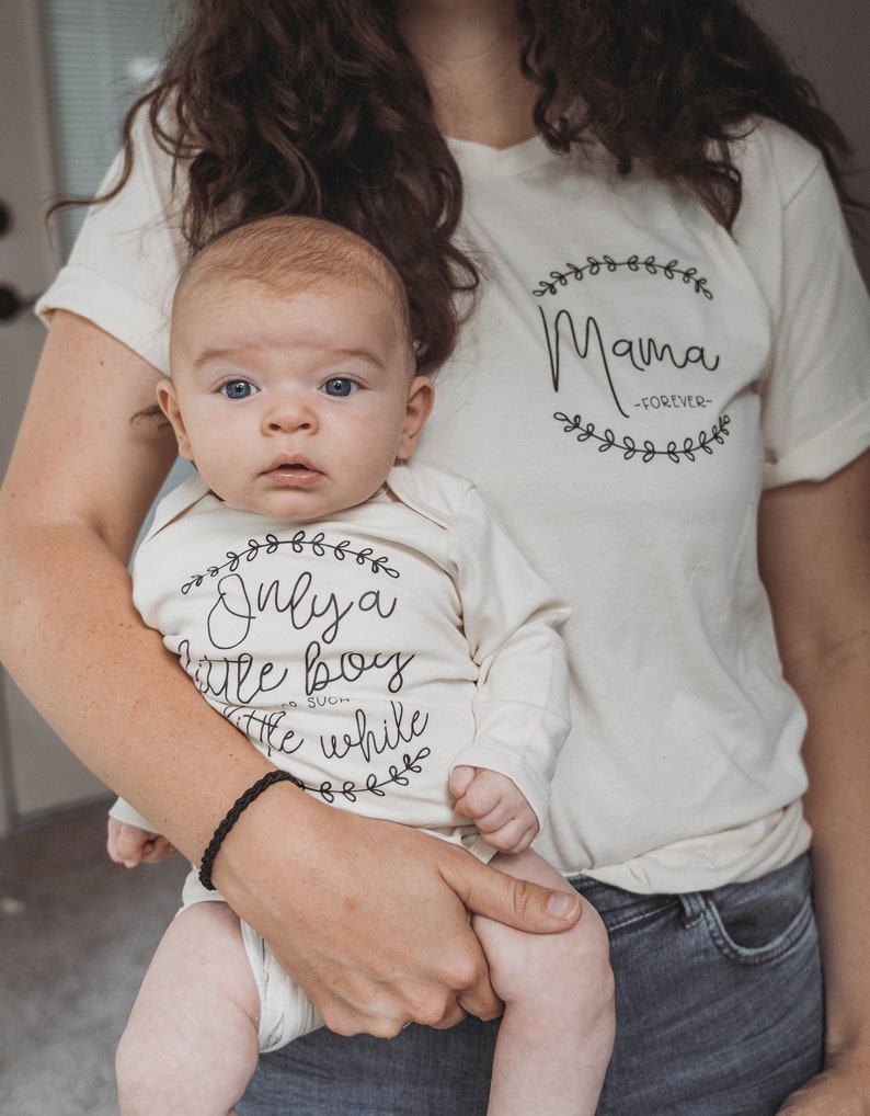 Pode incluir: Uma mulher segurando um beb&ecirc; que est&aacute; vestindo um macac&atilde;o branco com o texto "Only a little boy for such a while" em uma coroa de flores preta. A mulher est&aacute; vestindo uma camiseta branca com o texto "Mama -FOREVER- " em uma coroa de flores preta.