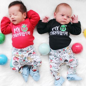 May include: Two babies wearing matching Christmas outfits with the text "My 1st Christmas" on their onesies. The babies are wearing white pants with a Christmas pattern and blue moccasins. The babies are surrounded by colorful Christmas ornaments.