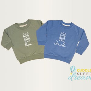 May include: Two green and blue children's sweatshirts with white text and a graphic of three candles. The green sweatshirt says "Three" and the blue sweatshirt says "Jack".