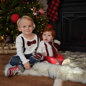 May include: Two young boys wearing matching red and black bow ties and suspenders. The older boy is wearing blue jeans and red sneakers. The younger boy is wearing red pants and is sitting on a white furry rug. They are both smiling at the camera.