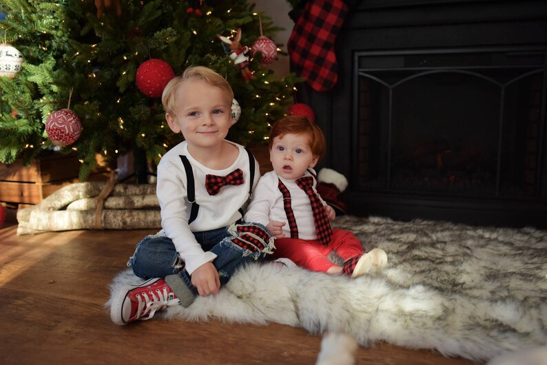 May include: Two young boys wearing matching red and black bow ties and suspenders. The older boy is wearing blue jeans and red sneakers. The younger boy is wearing red pants and is sitting on a white furry rug. They are both smiling at the camera.